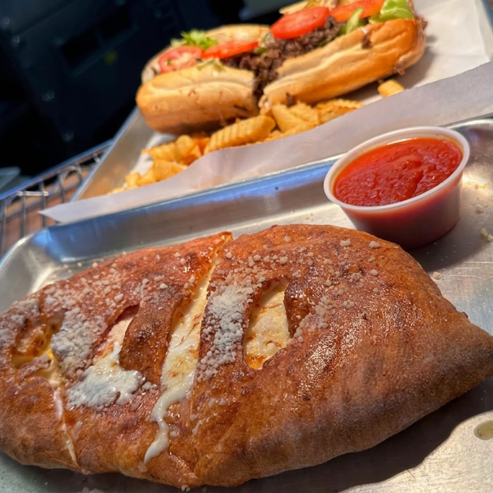 Baked calzone topped with grated cheese and served with marinara sauce, alongside hot dogs and fries on metal trays.