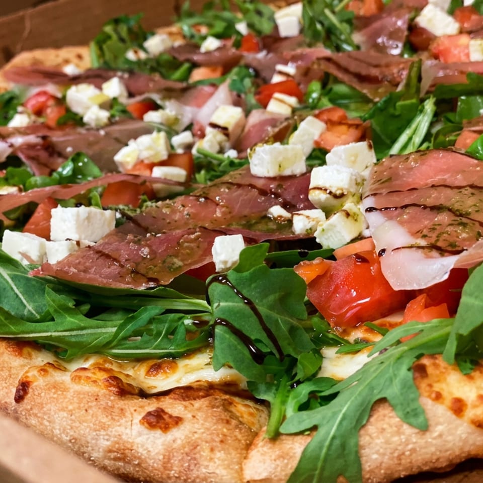 Close-up of a rectangular pizza topped with arugula, prosciutto, feta cheese, tomatoes, and balsamic glaze.